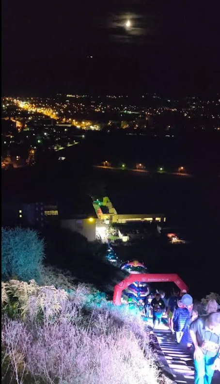 Tekači ponoči na osvetljenih stopnicah Kalvarije, pod njimi napihljiv startni lok in luči Maribora.